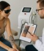 Een tatoeageverwijderingsspecialist voert een intakegesprek en huidanalyse uit met een cliënt die een bloemen-tatoeage op haar bovenarm heeft, in een schone medische ruimte met laserapparatuur op de achtergrond.