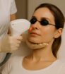 Vrouw ondergaat een laserbehandeling in een kliniek, met oogbescherming en een professionele laserhandstuk op haar huid gericht.