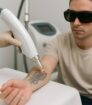 Laserbehandeling van een zwart/grijze tattoeage op de onderarm; patiënt met beschermbril in kliniek.
